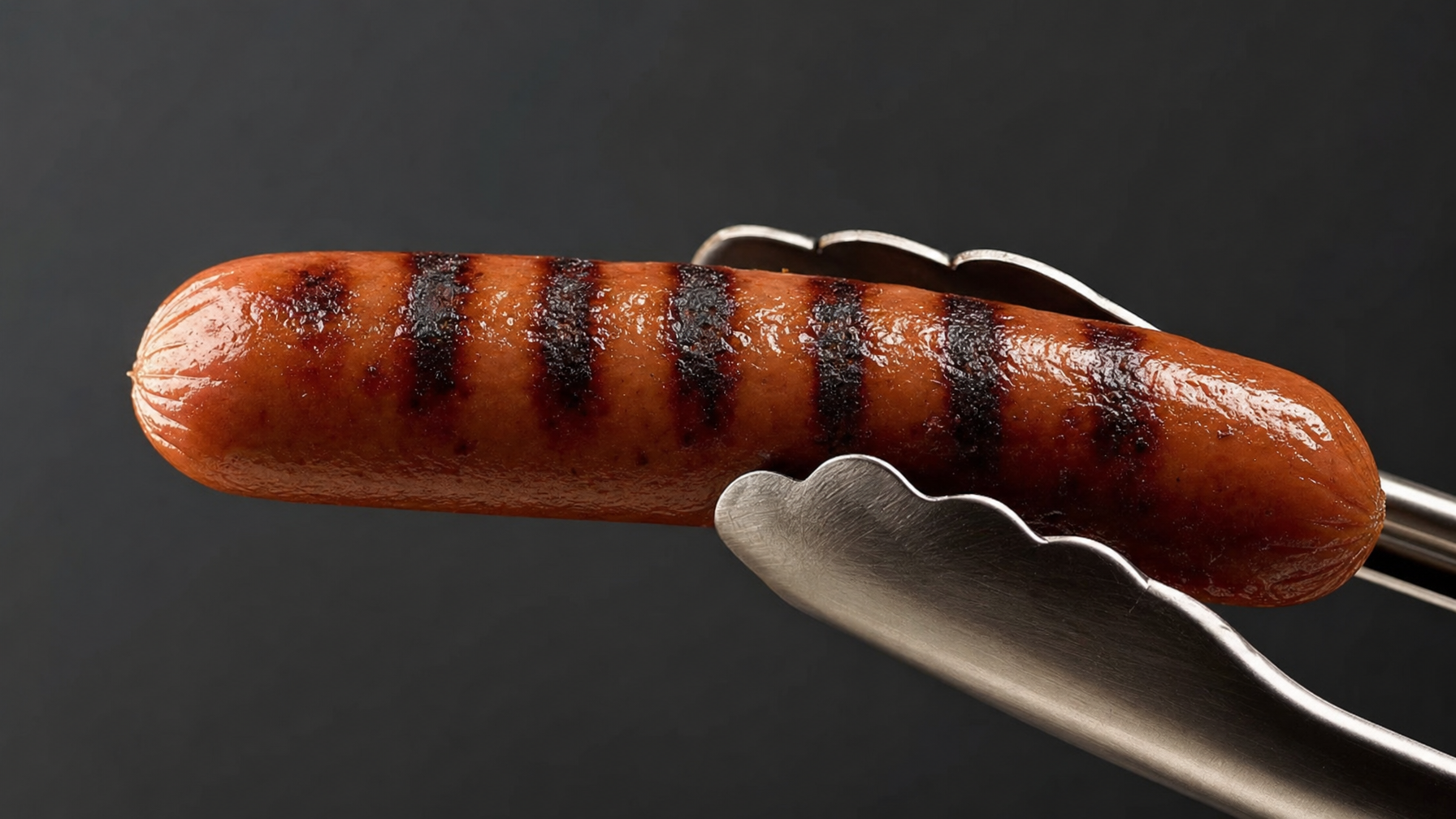 A grilled hot dog with dark grill marks is held in metal tongs against a dark background.